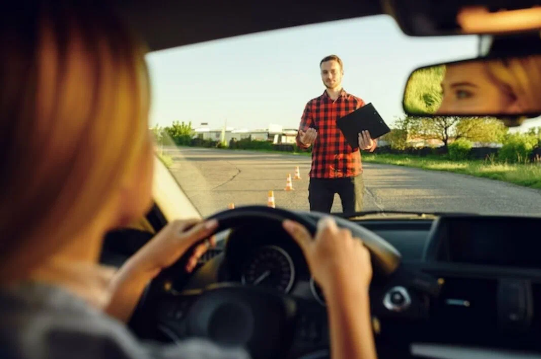 View from the driver's seat of a car looking at a driving instructor with a clipboard and traffic cones
